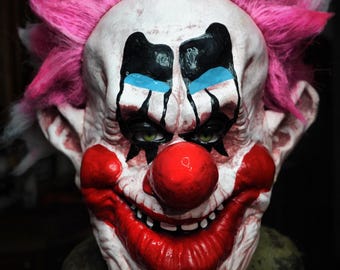 May include: A creepy clown mask with a white face, black eye makeup, and a red nose and mouth. The mask has pink spiky hair and green eyes. The clown mask is a Halloween costume.