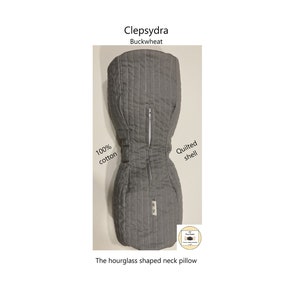 May include: Gray hourglass-shaped neck pillow with a zipper down the back. The pillow is made of 100% cotton and has a quilted shell. The text "Clepsydra Buckwheat" is printed on the top of the pillow.
