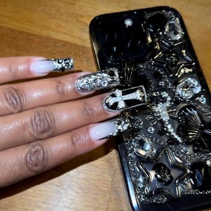 May include: Black phone case with a silver and black design featuring a cross, skulls, and stars. The case is decorated with rhinestones. A hand with long, white nails decorated with silver and black designs is holding the phone case.