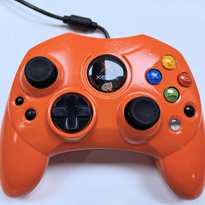 Könnte beinhalten: Orangefarbener Xbox-Controller mit schwarzen Knöpfen und einem schwarzen kreuzförmigen Steuerkreuz. Der Controller hat farbige Aktionstasten und ein Mountain Dew-Logo. Ein schwarzes Kabel erstreckt sich von oben.