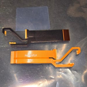May include: Two flexible circuit boards. One is black with a textured surface and a copper-colored section. The other is orange with the text "XeriamFLX" and a series of parallel lines. Both have angled, hook-shaped ends.