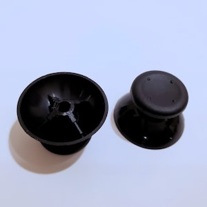 Puede incluir: Dos palancas de pulgar de repuesto negras para un mando de videojuegos. Una palanca de pulgar es plana en la parte superior y la otra tiene una parte superior cóncava.