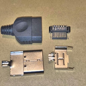 May include: An assortment of black and silver electronic components, including a connector with a ribbed end, a small rectangular piece with pins, and two silver metal pieces with tabs and slots. The components are arranged on a neutral-toned surface.