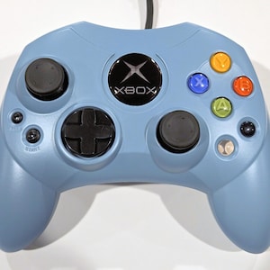 以下が含まれることがあります： 黒いアナログスティックと黒い十字キーを備えた、水色のXboxコントローラー。 コントローラーにはカラーボタンとXboxのロゴが付いています。 「XBOX」の文字がコントローラーに印刷されています。