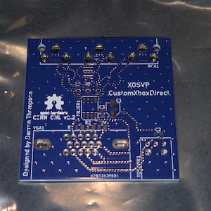 Może przedstawiać: Niebieska płytka drukowana z miedzianymi ścieżkami i elementami elektronicznymi. Tekst na płytce to "XOSVP CustomXboxDirect," "CERN CHL v2.2," i "Designed by Darren Thompson." Posiada wiele punktów połączeń.
