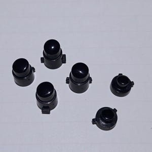May include: A collection of six black plastic components, possibly parts of a larger mechanism. The pieces have a rounded top and a cylindrical base with small protrusions. The items are arranged on a white surface.