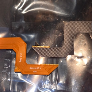 May include: Two flexible circuit boards. One is orange with the text "v1.5 Only" and "XeniumFLX v3.0". The other is gray with a crosshatch pattern. Both have gold-colored connectors.