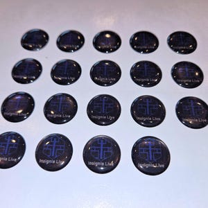 May include: Twenty dark, round, glass-like objects with a blue cross design and the text "Insignia Live". The objects are arranged on a white surface. The design is a dark blue color.