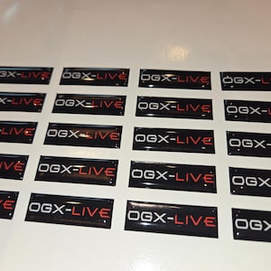 May include: A collection of black rectangular stickers with the text "OGX-LIVE" in white and red. The stickers are arranged on a white surface, with a glossy finish.