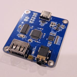 May include: A small, rectangular blue circuit board with various electronic components, including chips, ports, and connectors. It features a USB port, a USB-C port, and a headphone jack. The board is designed for electronic projects.