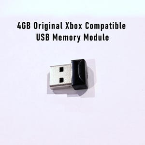 May include: A USB memory module for the original Xbox, with a silver USB connector and a black cap. The text "4GB Original Xbox Compatible USB Memory Module" is printed above the device.