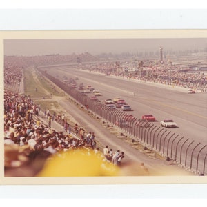 Op de afbeelding: Een vintage foto toont een autorace. Verschillende auto's in verschillende kleuren racen op een circuit. Toeschouwers vullen de tribunes aan beide zijden van de baan, wat een levendige sfeer creëert. De foto is genomen vanuit een iets verhoogd perspectief.