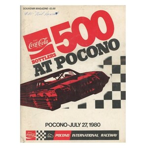 Könnte beinhalten: Souvenir-Magazin-Cover für das Pocono 500-Rennen 1980. Das Cover zeigt ein rotes Rennauto mit Bewegungsunschärfe, das Coca-Cola-Logo und den Text "500 at Pocono". Das Datum "27. Juli 1980" ist ebenfalls angegeben.