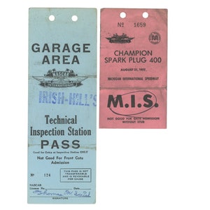 May include: Two vintage racing passes. One is light blue with "GARAGE AREA" and "NASCAR" text. The other is pink, labelled "CHAMPION SPARK PLUG 400" and "M.I.S.". Both have holes for hanging and are from the Michigan International Speedway.