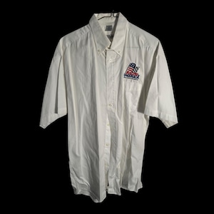 May include: White short-sleeve button-down shirt with a small pocket on the left chest. The shirt features a Racing America logo with a flag design in red, white, and blue. The shirt is hanging against a black background.