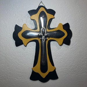 May include: A black and gold wooden cross with a silver fleur-de-lis design. The cross is hanging on a white wall.