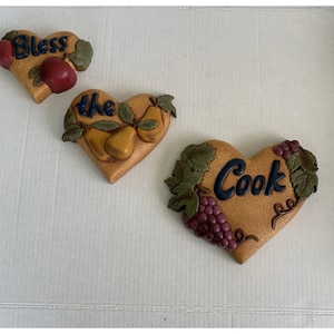 Può includere: Tre placche decorative per la cucina a forma di cuore con uno stile rustico e vintage. Le placche presentano una mela rossa, pere e uva, e le parole "Bless the Cook".