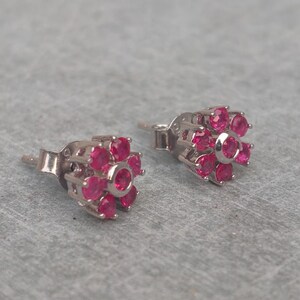 May include: A pair of silver-toned stud earrings, each featuring a flower design. The flowers are composed of small, round, bright pink gemstones. The earrings are set against a light gray background.