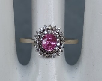 May include: A gold ring featuring a pink oval gemstone surrounded by small white gemstones. The ring is set on a white surface, showcasing the intricate details of the jewelry. The ring's band is a simple gold color.