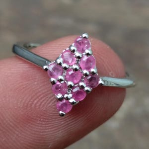 Könnte beinhalten: Ein silberner Ring mit einem Cluster aus kleinen, runden, rosa Edelsteinen, die in einer dreieckigen Form angeordnet sind. Der Ring wird von einem Finger gehalten, der das polierte Metallband und die leuchtende Farbe der Steine zeigt.