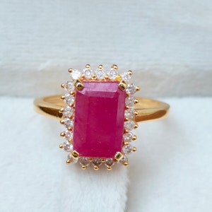 May include: A gold ring featuring a large, rectangular, ruby-colored gemstone. The ruby is surrounded by a halo of smaller, clear gemstones. The ring is set against a white, textured background.