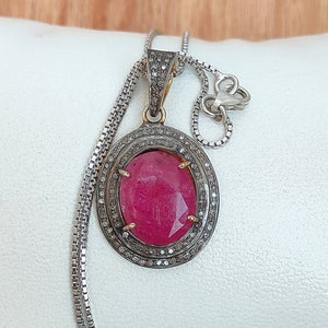 Op de afbeelding: Een zilveren ketting met een hanger met een grote ovale robijn, omringd door een halo van kleinere diamanten.