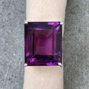 May include: A large, square-cut amethyst gemstone ring. The deep purple stone is set in a silver band. The ring is displayed on a white, textured surface against a gray background.