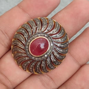 Può includere: Una spilla circolare con una pietra centrale rossa ovale a cabochon circondata da un bordo di piccole pietre incastonate. La sezione esterna è composta da segmenti metallici curvi e radianti accentuati da minuscole pietre scintillanti.