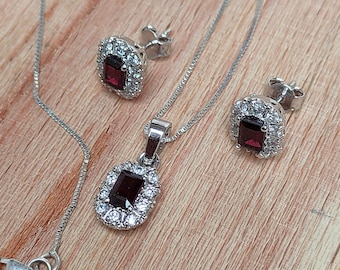 May include: A silver necklace and earrings set featuring a red gemstone surrounded by clear cubic zirconia stones.