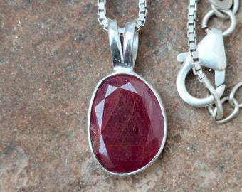 Op de afbeelding: Een zilveren ketting met een hanger met een rode ovale edelsteen.