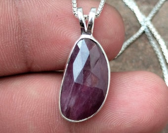 Op de afbeelding: Een gefacetteerde, ovaalvormige robijn hanger met een zilveren zetting hangt aan een fijne zilveren ketting. De dieprode edelsteen heeft meerdere facetten. De hanger is een sieraad.
