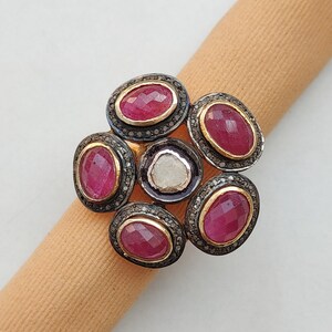 May include: A gold-toned ring featuring a cluster of oval-shaped, deep red gemstones. Each gemstone is surrounded by a halo of small, clear stones. The ring's design resembles a flower, with a central, irregularly shaped, white stone.