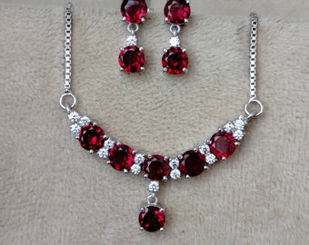 May include: A jewellery set comprising a necklace and earrings. The necklace and earrings have a silver chain and setting. The necklace features a curved design with multiple large, round, deep red gemstones and smaller white gemstones. The earrings have a similar design.