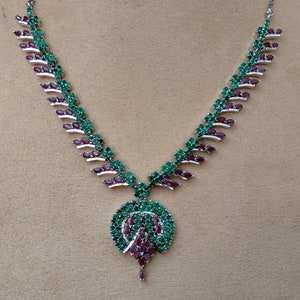 May include: A silver-coloured jewellery set comprising a necklace and matching earrings. The necklace features a leaf-like design with alternating green and burgundy-coloured stones. The earrings have a similar design, with a cluster of burgundy stones.