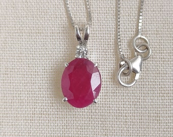 May include: A silver necklace featuring a large, oval-cut ruby pendant. The ruby is a deep red color and is accented by a small diamond above it. The chain is a delicate silver box chain.