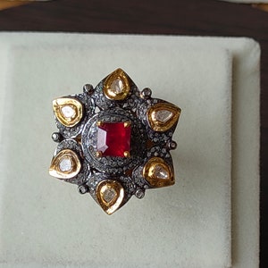 May include: A detailed, star-shaped ring featuring a central, square-cut red gemstone. The ring has a dark metal setting with small, clear accent stones and gold-colored teardrop-shaped elements. The ring is set on a white surface.