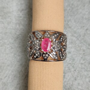 Puede incluir: Anillo plateado con una gema rosa ovalada brillante en el centro. El anillo presenta un diseño floral con pequeñas piedras transparentes. Se muestra sobre un objeto cilíndrico beige sobre un fondo gris.