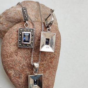 Peut inclure: Un ensemble collier et boucles d'oreilles en argent avec une pierre pr&eacute;cieuse bleue et des accents de marcassite. Le pendentif et les boucles d'oreilles sont de forme carr&eacute;e avec un bord biseaut&eacute;. Le dos du pendentif et des boucles d'oreilles est estampill&eacute; "925".