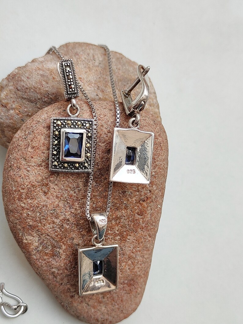 Peut inclure: Un ensemble collier et boucles d'oreilles en argent avec une pierre pr&eacute;cieuse bleue et des accents de marcassite. Le pendentif et les boucles d'oreilles sont de forme carr&eacute;e avec un bord biseaut&eacute;. Le dos du pendentif et des boucles d'oreilles est estampill&eacute; "925".