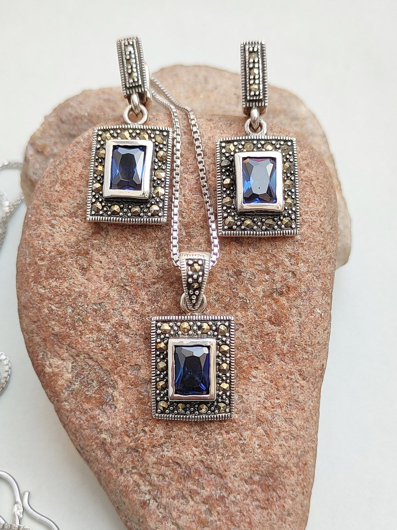 Peut inclure: Un ensemble de collier et boucles d'oreilles en argent avec une pierre pr&eacute;cieuse bleue et des accents de marcassite. Le pendentif et les boucles d'oreilles sont de forme carr&eacute;e avec une pierre pr&eacute;cieuse bleue rectangulaire au centre.