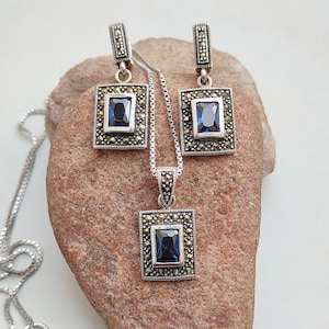 Peut inclure: Un collier et des boucles d'oreilles en argent avec une pierre pr&eacute;cieuse bleue et des accents de marcassite. Le pendentif et les boucles d'oreilles sont de forme carr&eacute;e avec un sertissage &agrave; b&eacute;li&egrave;re.
