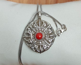 Puede incluir: Un collar de plata con un colgante de diseño detallado y ornamentado, con una piedra preciosa roja en el centro. El collar está sobre una superficie blanca.