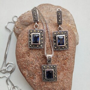 Peut inclure: Un ensemble de collier et boucles d'oreilles en argent avec une pierre pr&eacute;cieuse bleu fonc&eacute; et des accents de marcassite. Le pendentif et les boucles d'oreilles sont de forme carr&eacute;e avec une cha&icirc;ne en argent.