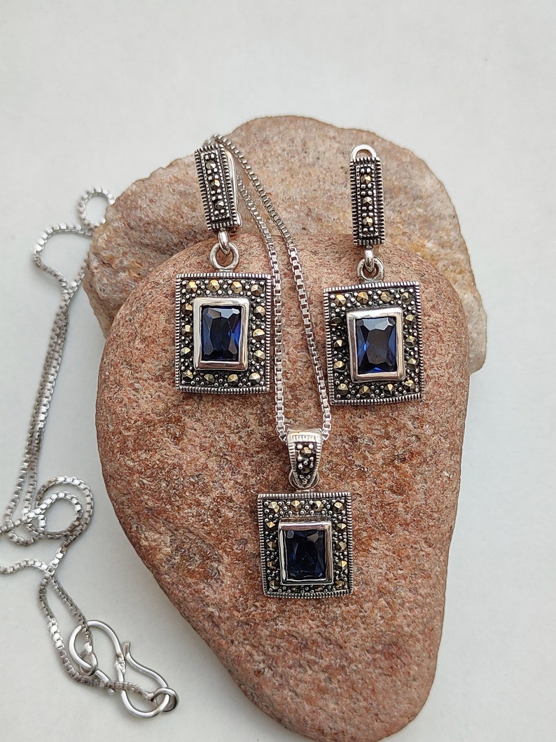 Peut inclure: Un ensemble de collier et boucles d'oreilles en argent avec une pierre pr&eacute;cieuse bleu fonc&eacute; et des accents de marcassite. Le pendentif et les boucles d'oreilles sont de forme carr&eacute;e avec une cha&icirc;ne en argent.