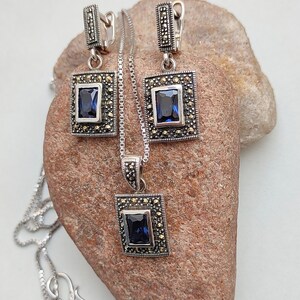 Peut inclure: Un ensemble de collier et boucles d'oreilles en argent avec une pierre pr&eacute;cieuse bleue et des accents de marcassite. Le pendentif et les boucles d'oreilles pr&eacute;sentent un design carr&eacute; avec une pierre pr&eacute;cieuse bleue au centre.
