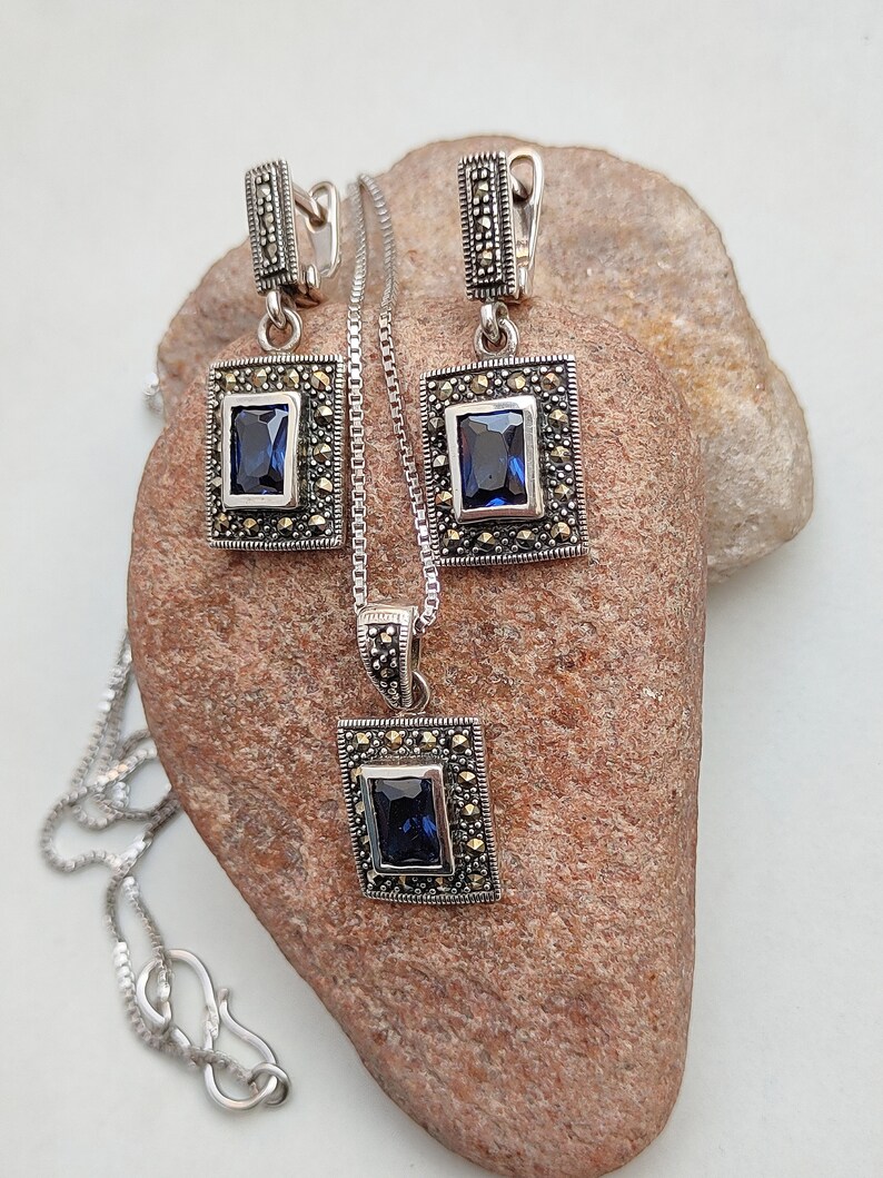 Peut inclure: Un ensemble de collier et boucles d'oreilles en argent avec une pierre pr&eacute;cieuse bleue et des accents de marcassite. Le pendentif et les boucles d'oreilles pr&eacute;sentent un design carr&eacute; avec une pierre pr&eacute;cieuse bleue au centre.