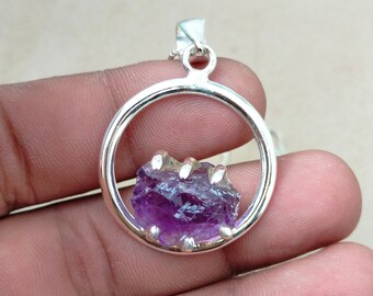 May include: A silver pendant with a raw amethyst gemstone set in a circular frame. The amethyst is a deep purple color and has a rough, natural texture.