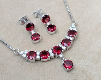 May include: A silver necklace and earrings set with a red gemstone pendant and red gemstone accents. The necklace has a silver chain and a curved design with small clear stones.