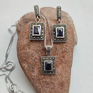Peut inclure: Un ensemble de collier et boucles d'oreilles en argent avec un pendentif en pierre bleue et des boucles d'oreilles assorties. Le pendentif et les boucles d'oreilles sont entour&eacute;s de petites pierres scintillantes.