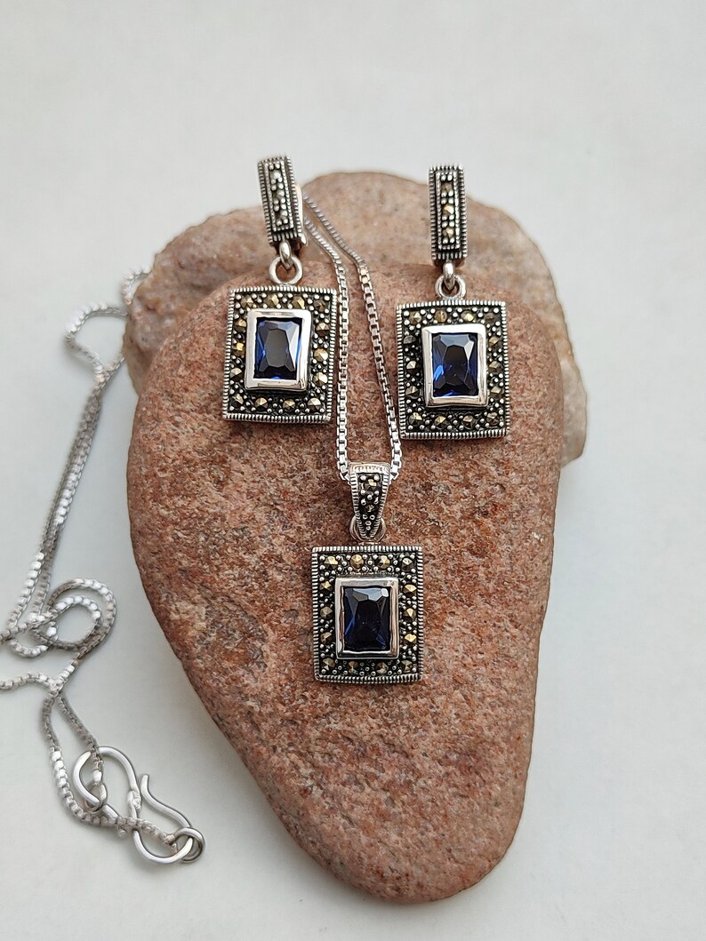 Peut inclure: Un ensemble de collier et boucles d'oreilles en argent avec un pendentif en pierre bleue et des boucles d'oreilles assorties. Le pendentif et les boucles d'oreilles sont entour&eacute;s de petites pierres scintillantes.